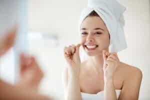 Young woman with hair in a towel flossing her teeth while looking in a bathroom mirror