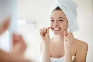 Young woman with hair in a towel flossing her teeth while looking in a bathroom mirror