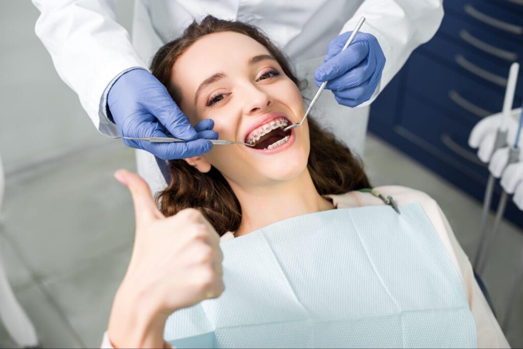 Smiling female patient with braces giving a thumbs up while a dentist performs an exam